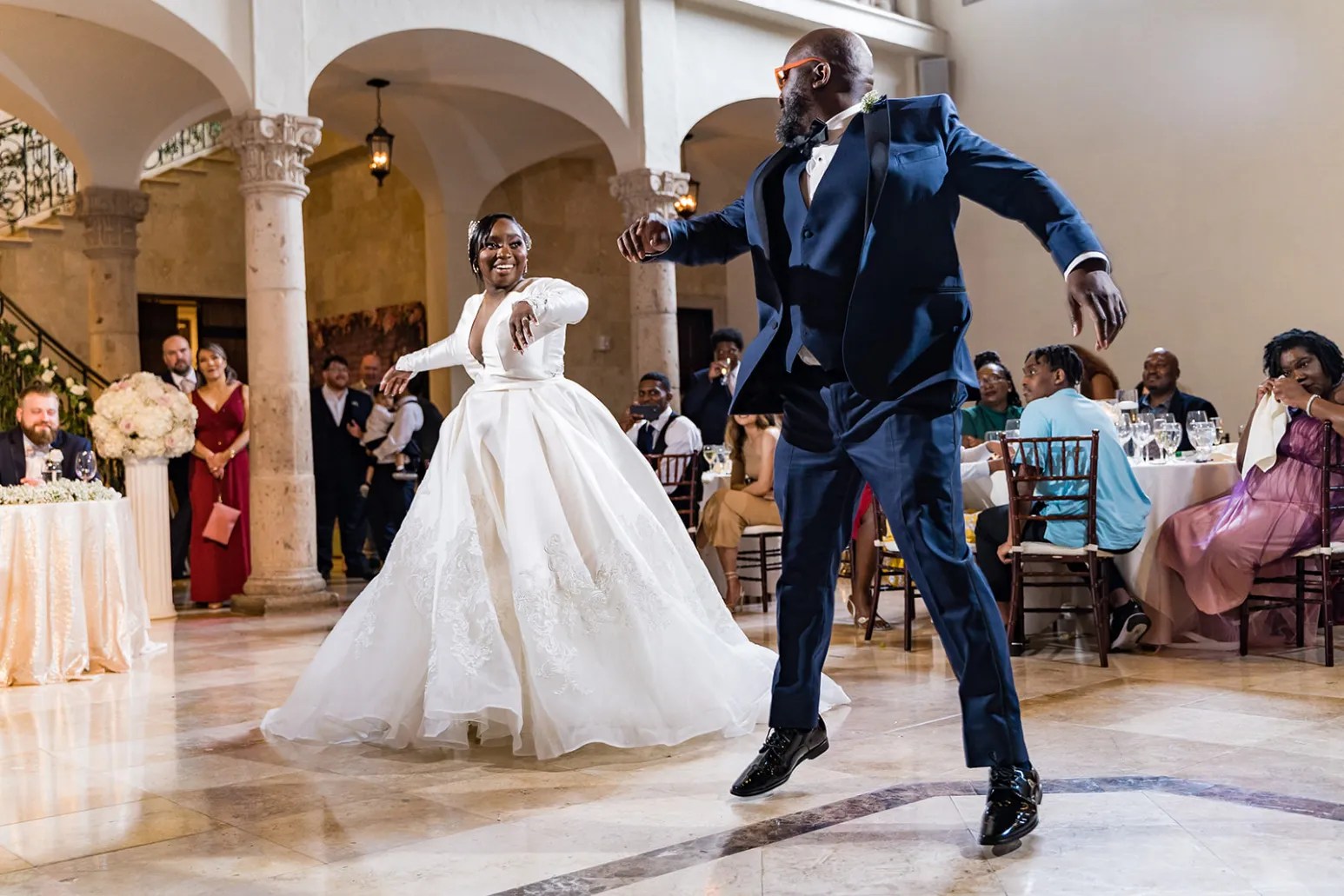 Ashley and Benjamin Bride and groom energetically dancing in elegant wedding attire during the reception, surrounded by guests in a beautifully decorated venue. Wedding Photography Services Capture Every Moment.