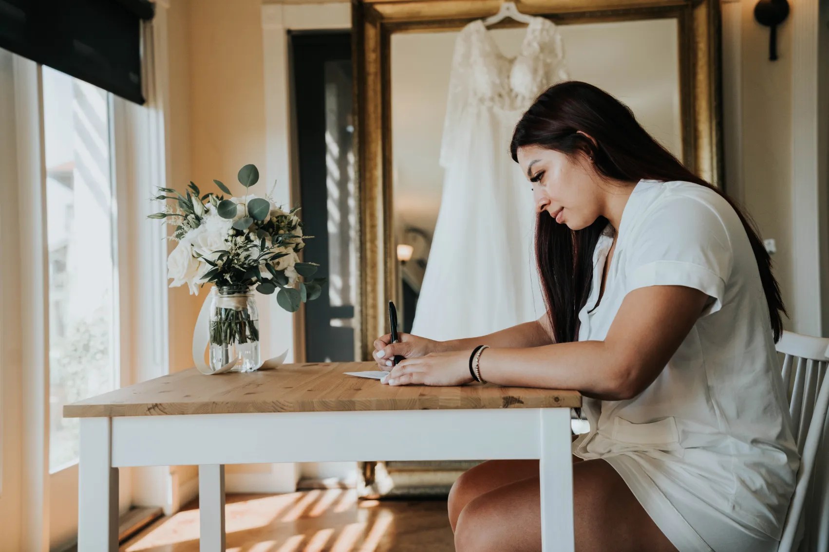 bride writing vows Bride writing at a table with a bouquet of flowers and a wedding dress in the background, capturing wedding preparations.