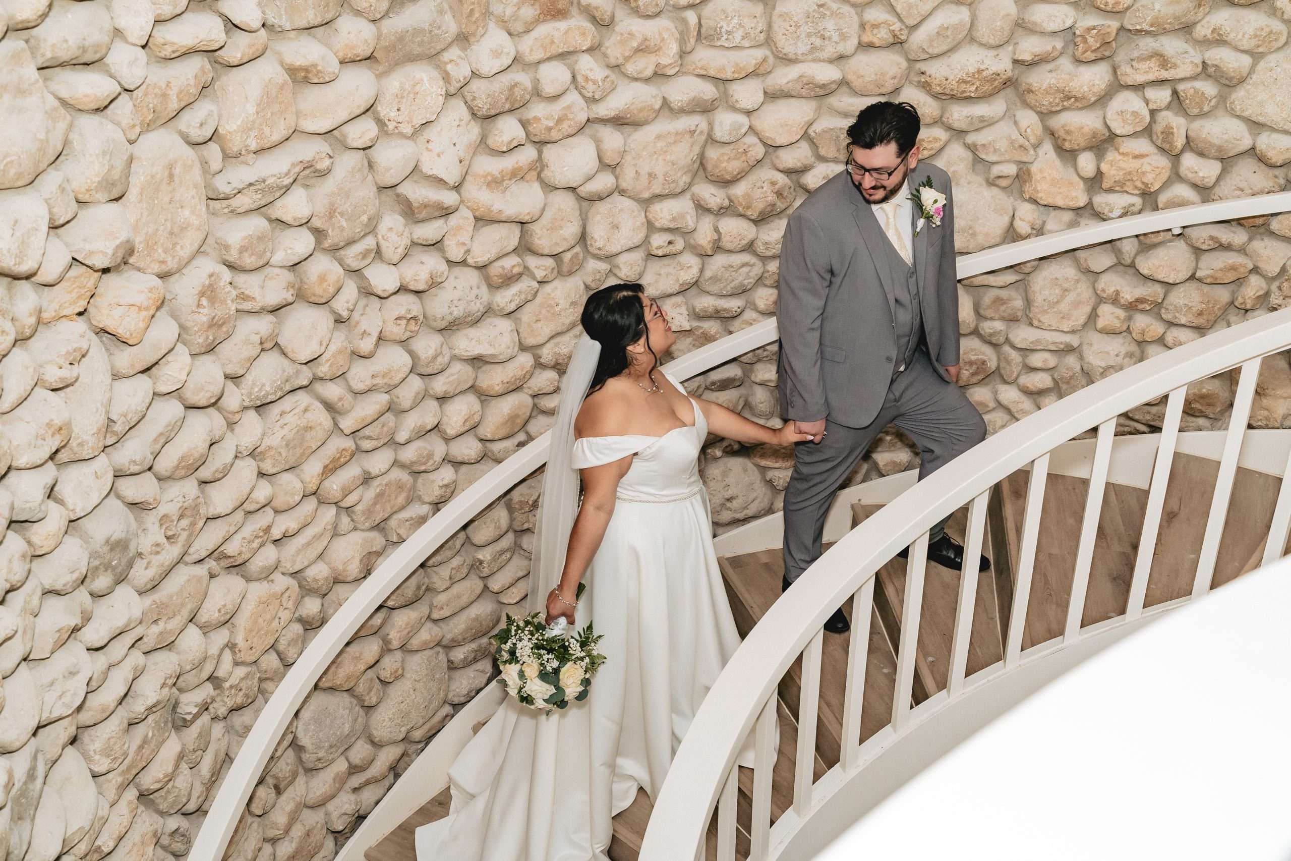 Bride and groom holding hands on white staircase against textured stone wall at indoor wedding venue