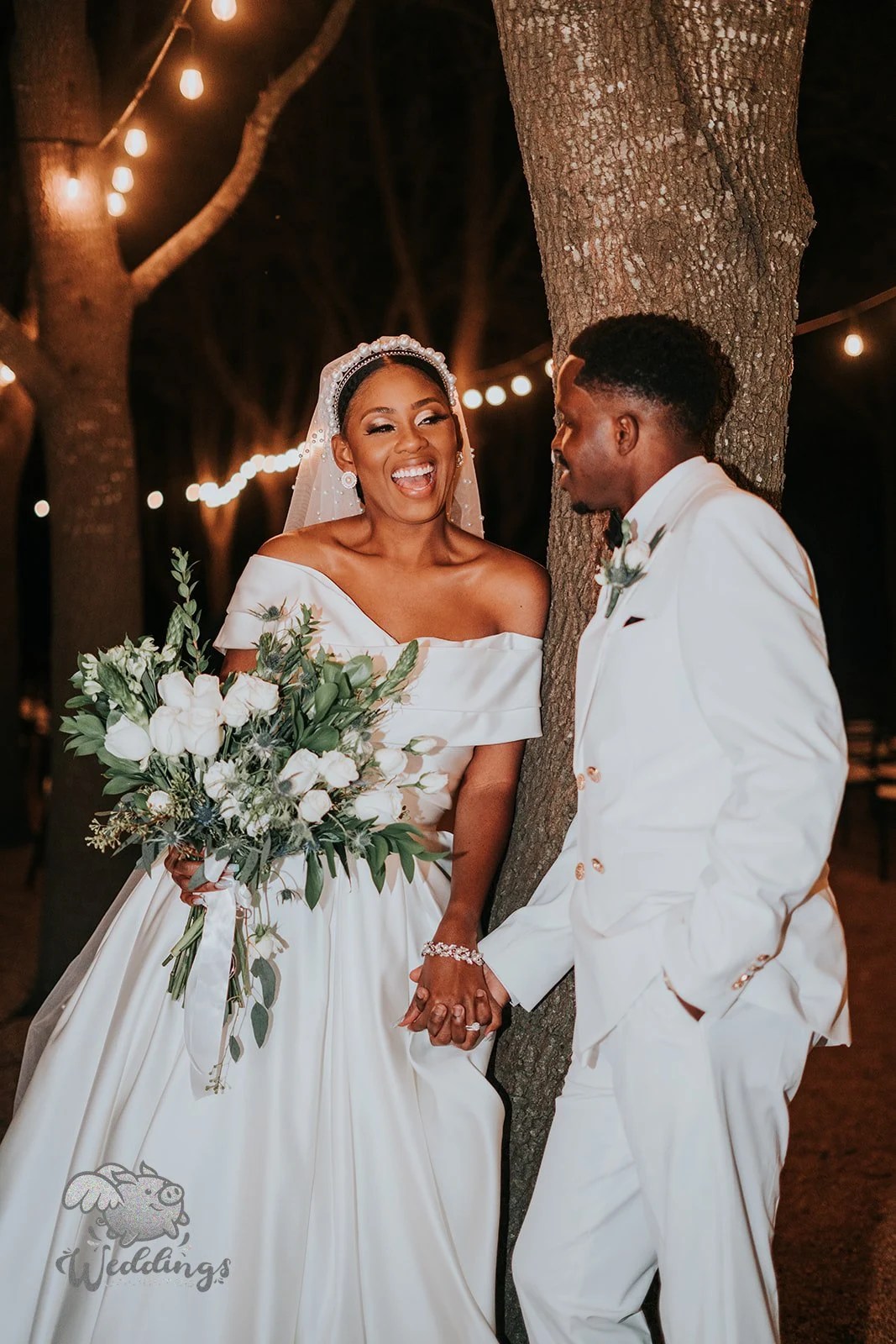 Bride laughing in an off shoulder satin gown with pearl headband holding a white rose bouquet under warm string lights at night