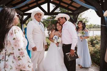 Bride in white dress and groom in white suit with cowboy hat standing under wooden pavilion with wedding guests
