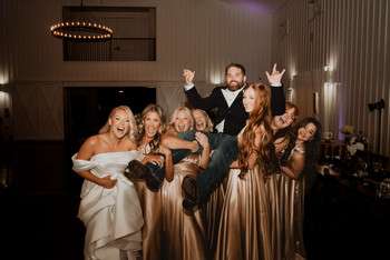 Wedding party celebrating with raised arms in rustic reception venue with string lights and fireplace