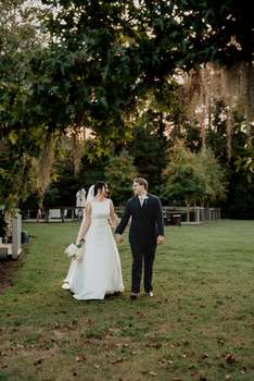 Bride in white gown and groom in navy suit walk hand in hand across green lawn with trees and chandelier overhead