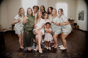 Bride in white dress surrounded by bridesmaids in mismatched neutral dresses and young flower girl indoors