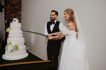 Bride and groom cutting white tiered wedding cake with sword at indoor reception