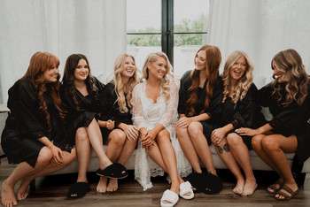 Bride in white lace dress and white slippers sits with six bridesmaids in black robes on bench by window