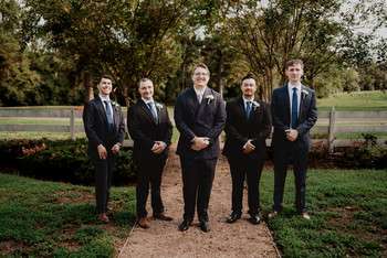 Groom with four groomsmen in black suits standing on pathway with trees and lawn in background