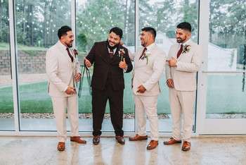 Groom in black suit with three groomsmen in beige suits standing by floor-to-ceiling windows
