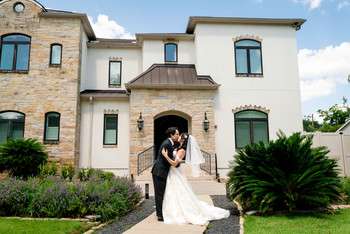 Bride and groom kissing under stone archway entrance of Mediterranean-style villa with manicured lawn