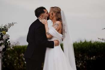 Bride and groom kiss during outdoor wedding ceremony with white floral arrangements in background