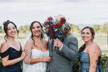 Bride with groom holding burgundy bouquet, flanked by two bridesmaids in dark green dresses on golf course
