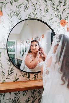 Bride applying makeup while looking in round mirror against botanical wallpaper with peach floral accents