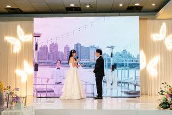 Bride and groom standing together during ceremony before white backdrop showing waterfront cityscape view