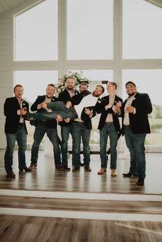Groom with groomsmen in casual attire holding small dogs indoors at venue with white walls and large windows