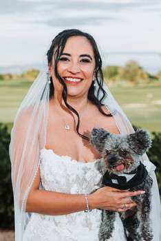 Bride in white lace wedding dress and veil smiling while holding small gray dog outdoors at golf course