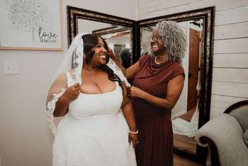 Bride in white strapless gown and veil smiles as woman in burgundy dress adjusts her veil during wedding preparation