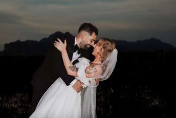 Groom in black tuxedo dips bride in white lace dress and veil for a kiss at sunset with mountains behind