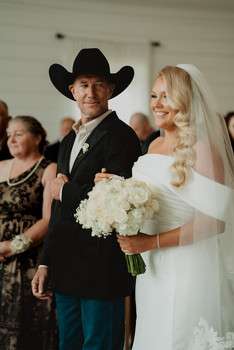 Bride in white off-shoulder dress with veil holds white bouquet beside groom in black cowboy hat and suit