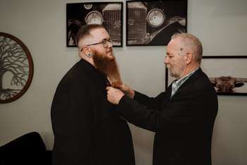 Older man in black suit helps groom with full beard adjust his tie before wedding ceremony