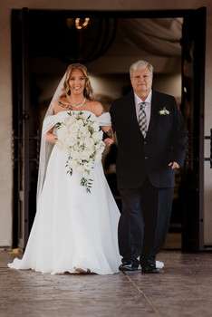 Bride in off-shoulder white gown holding white bouquet walks arm-in-arm with man in navy suit under covered walkway