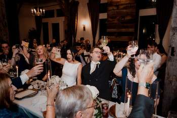 Wedding guests raise champagne glasses in toast at reception dinner in rustic barn venue with wooden walls
