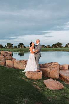 Bride and groom embracing on golf course lawn beside decorative boulders and pond under evening sky