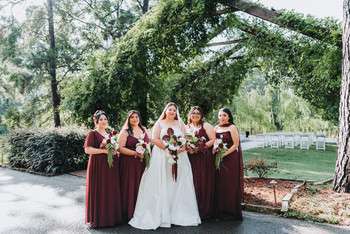 Bride in white gown with five bridesmaids in burgundy dresses holding bouquets under greenery arch outdoors
