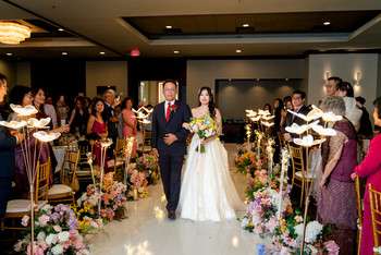 Bride and groom walk down aisle between flower arrangements and guests holding sparklers in reception hall