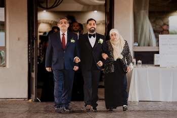 Groom in black tuxedo walks arm-in-arm with parents outside wedding venue