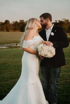 Bride and groom embrace on golf course lawn at sunset, bride holding white bouquet