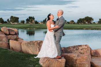 Bride and groom embrace on rock formation beside pond with golf course and mountain views in background