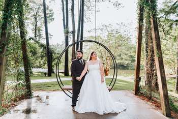 Bride and groom standing together inside circular metal arch decorated with greenery on outdoor ceremony platform