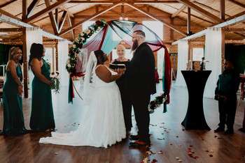 Bride and groom exchanging vows under draped arch in rustic wooden pavilion with wedding party and guests