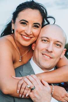 Bride embracing groom from behind, both smiling at camera, showing wedding rings against cloudy sky