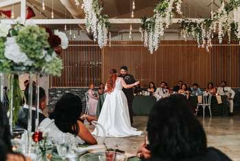 Bride and groom share first dance on wooden dance floor surrounded by seated wedding guests at reception