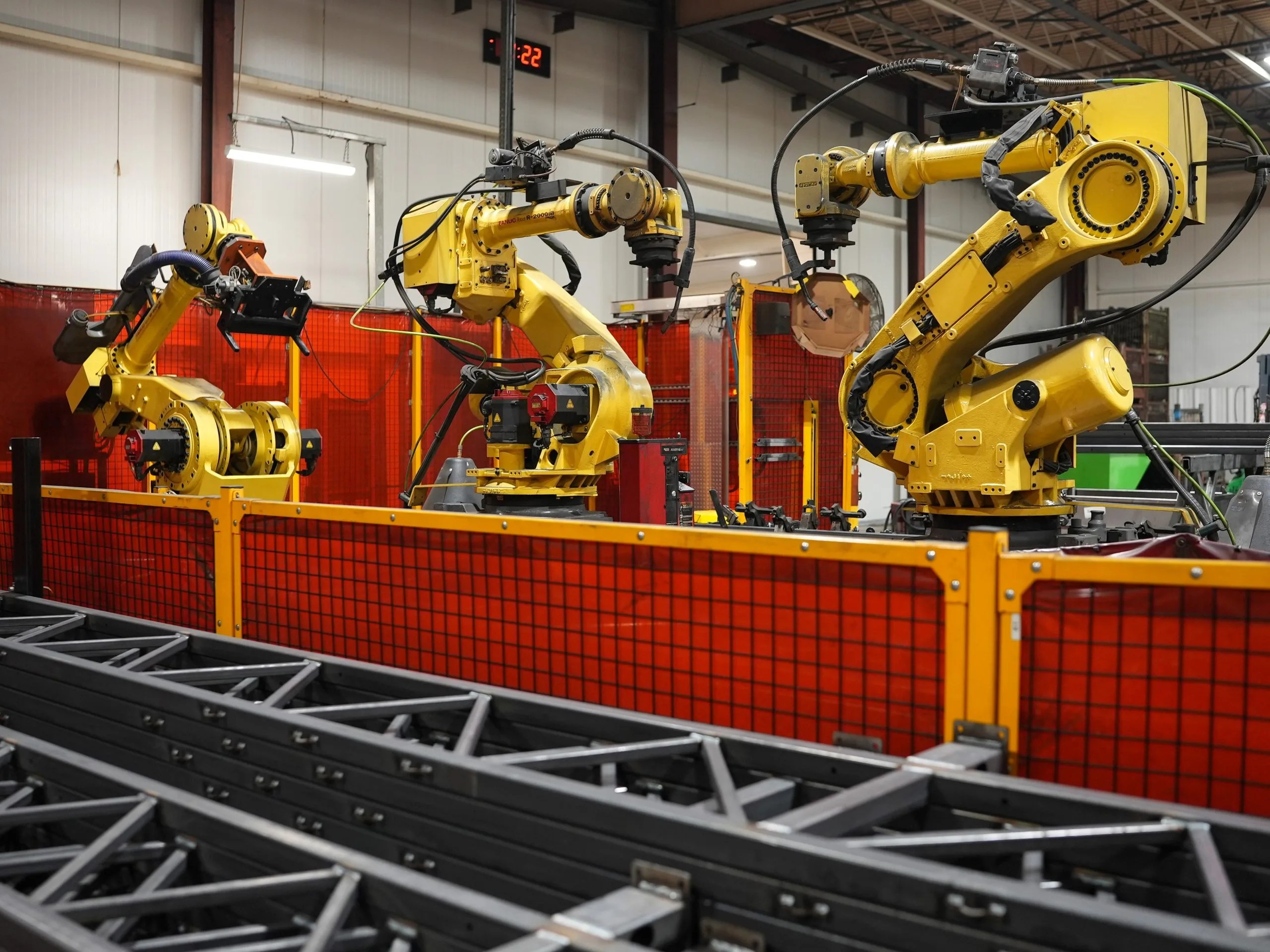 Three FANUC robotic welding arms in a fenced weld cell beside steel truss sections at the WeFab shop in Leamington, Ontario