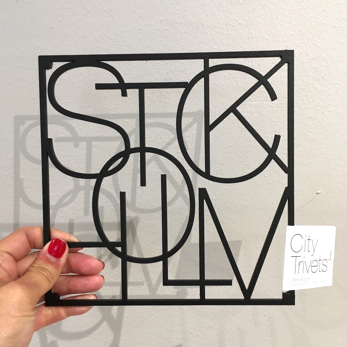 A metal trivet made up of letters, which spell out Stockholm. The trivet is square shaped and the letters overlap one another.