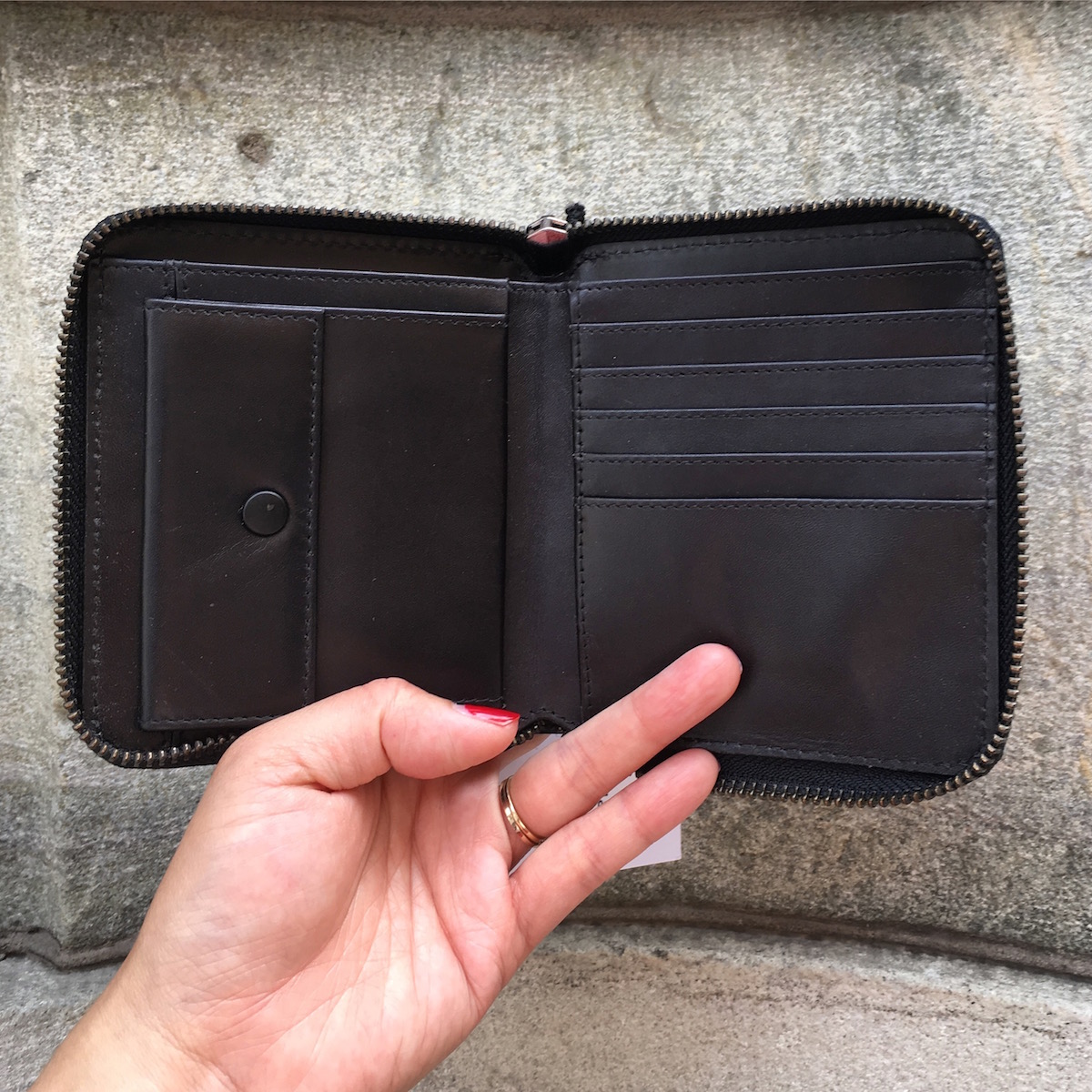 A hand holding open a zippered square wallet. The wallet has a coin pocket, five credit card slots, 2 additional slots, and a paper cash slot.