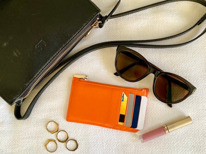 An orange card case with four credit card slots, along with a small black belt bag, sunglasses, lipstick, and some gold rings.