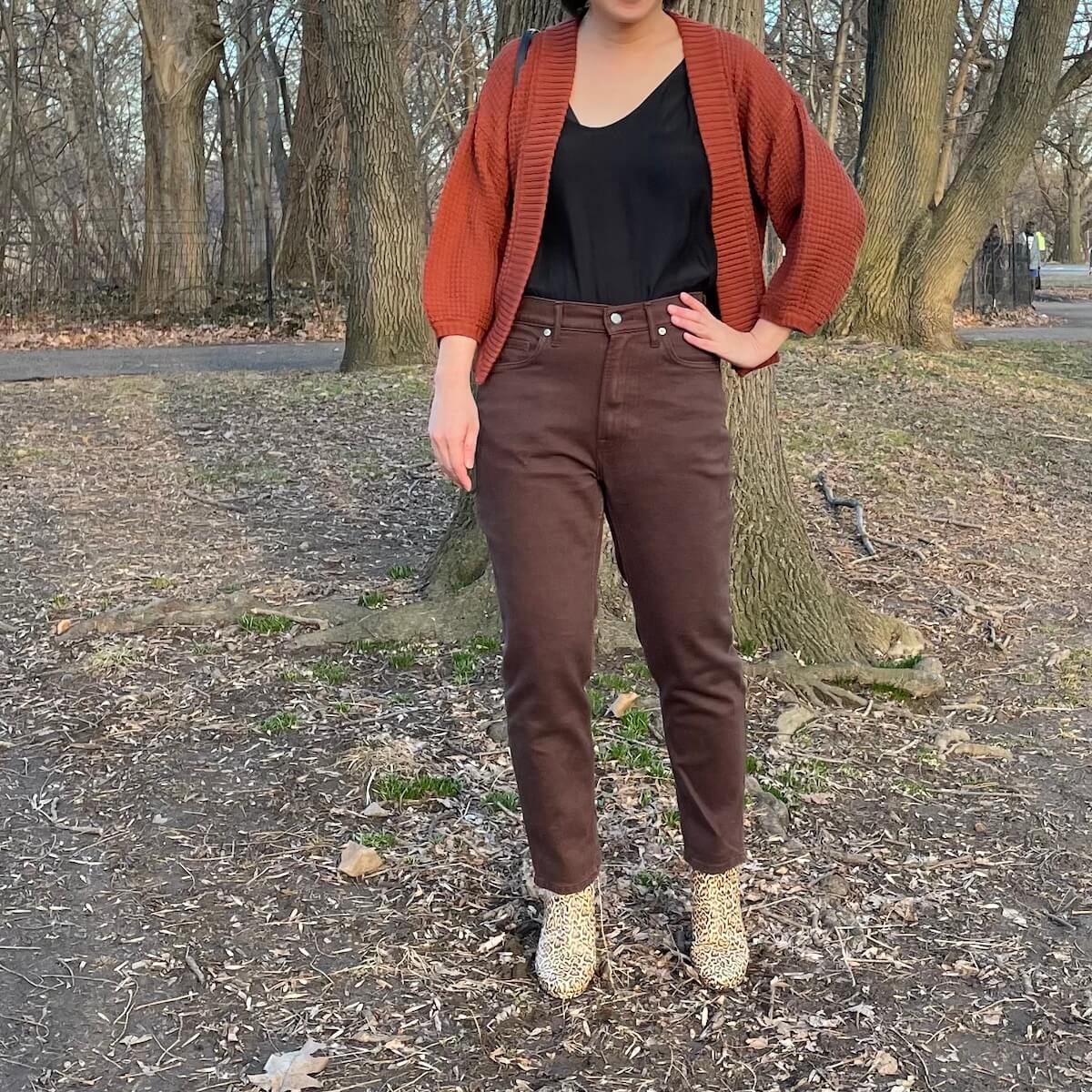 A person with dark hair, posing outside in brown Everlane Original Cheeky jean and a redish cardigan.