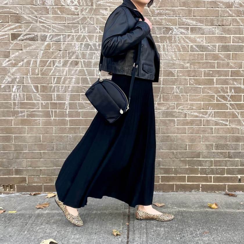 A person wearing a cropped black moto jacket and a black maxi skirt moves forward in front of a brick wall.