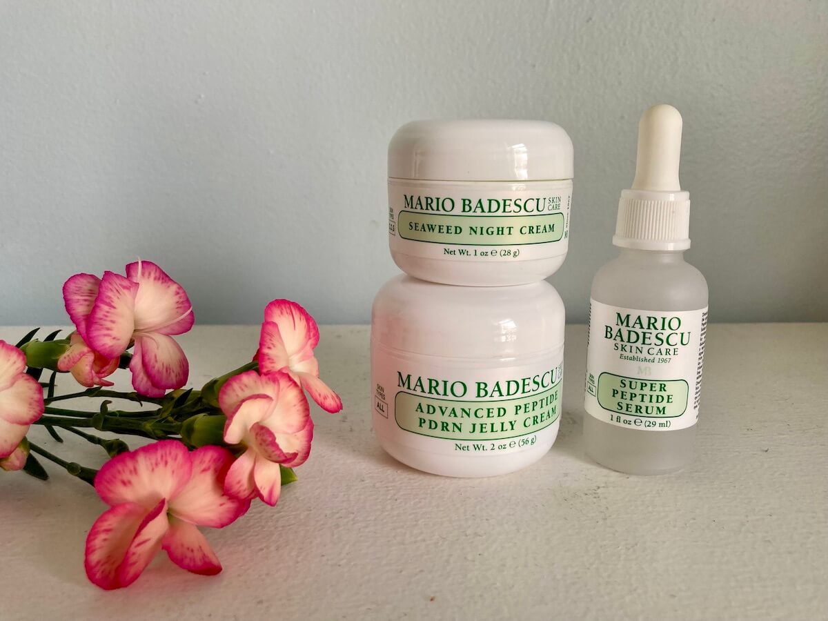 A jar of Mario Badescu Seaweed Night Cream stacked on a larger jar of Advanced Peptide PDRN Jelly Crea, next to a dropped jar of Super Peptide Serum. There are some red flowers to the left.