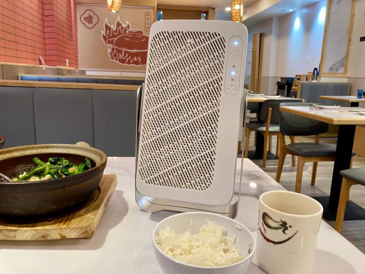 A white rectangular air purifier, the AirFanta 4Lite, on a table in a restaurant, with a clay pot, bowl of rice, and tea cup.