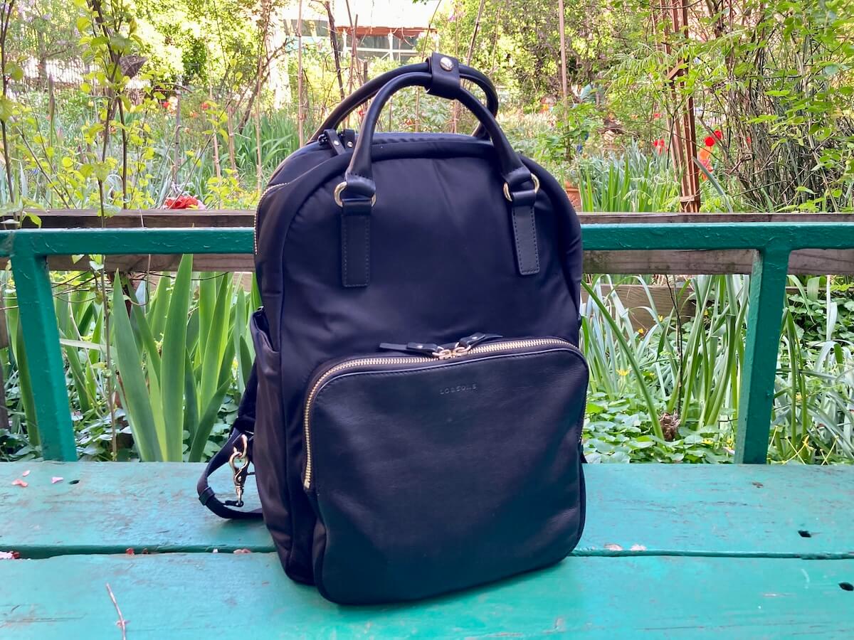 A black backpack made of nylon and leather on a green bench in a garden.
