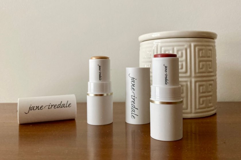 Two tubes of jane iredale products, which are open, on a wooden surface. There is a lidded white ceramic jar behind them.