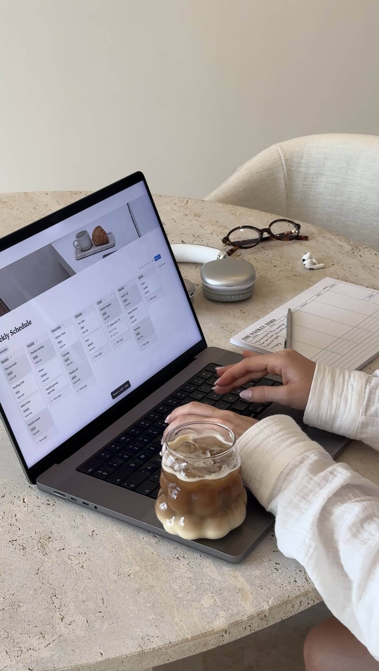 woman typing on a laptop with an iced coffee next to her on the table along with a paper schedule, headphones and glasses. Self-care routine. how to create a selfcare routine. creating a self-care ritual. how to build a routine for self-care.