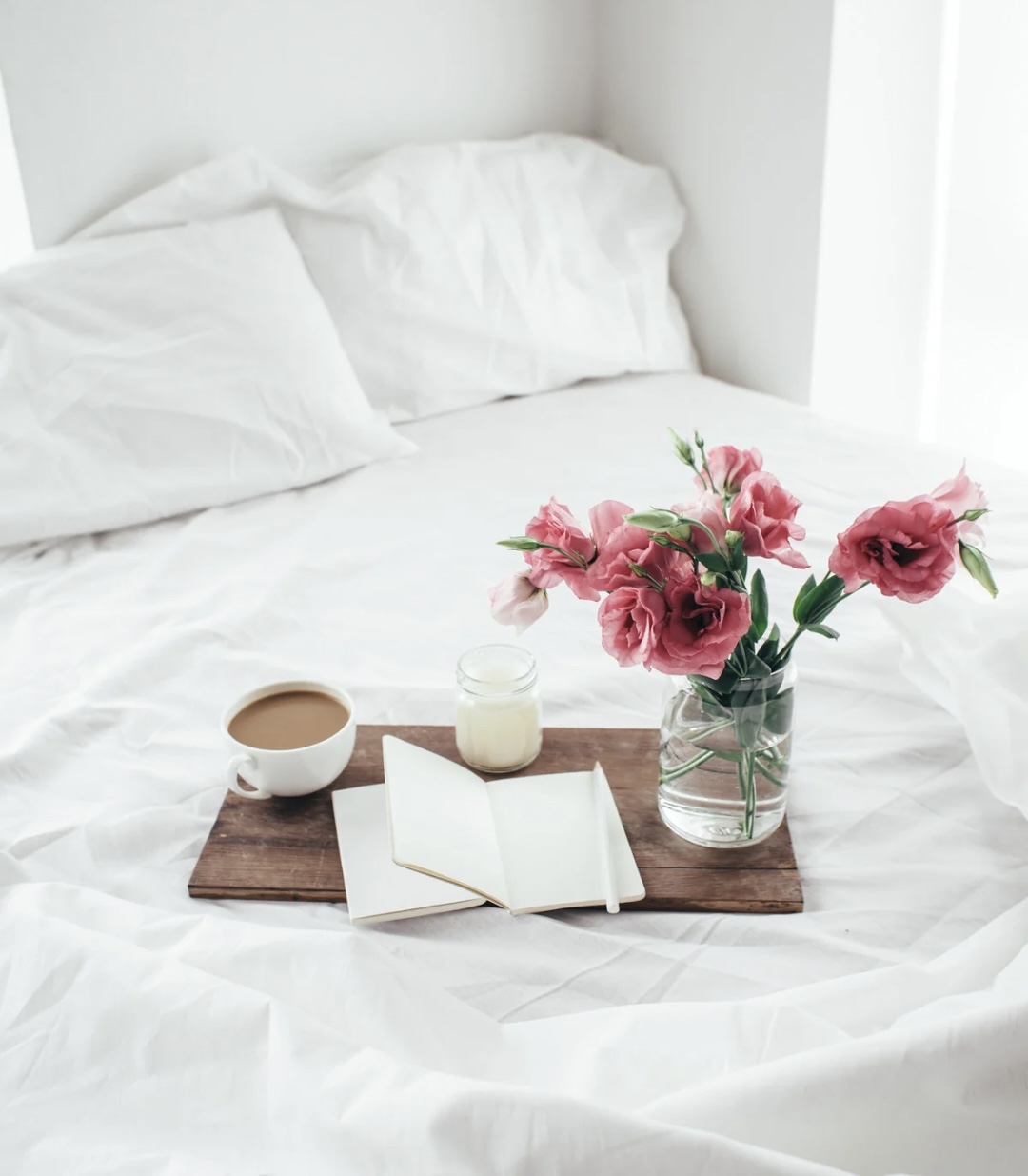 spring journal prompts. spring journaling ideas. journaling prompts for spring. questions to are in spring. seasonal journaling. spring self reflection. spring mindset reset. spring mental health ideas. A wooden board on a bed with white sheets with a notebook, candle, cup of coffee and a vase of pink flowers on top of it.