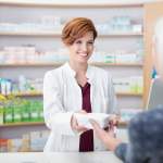 A friendly female pharmacist with auburn hair, dressed in a white coat over a maroon top, hands over a packaged item to an elderly customer, whose back is to the camera. They are at a pharmacy counter with shelves of medications visible in the background. The environment is clean and well-lit, indicative of a professional healthcare setting.