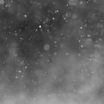 A grayscale abstract background with a multitude of defocused white specks scattered across a gradient from light to dark, resembling a gentle snowfall or concrete dust particles illuminated in soft light.