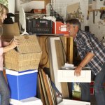 A man and woman organizing a cluttered garage, surrounded by various storage items like boxes, tools, and a wheelbarrow, indicating a cleaning or decluttering process in a domestic setting, possibly in spring time.